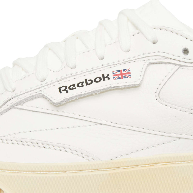 Close-up of a white Reebok sneaker, showing white laces, stitching, and the Reebok logo with a small Union Jack flag on the side. The sole is a light beige color.
