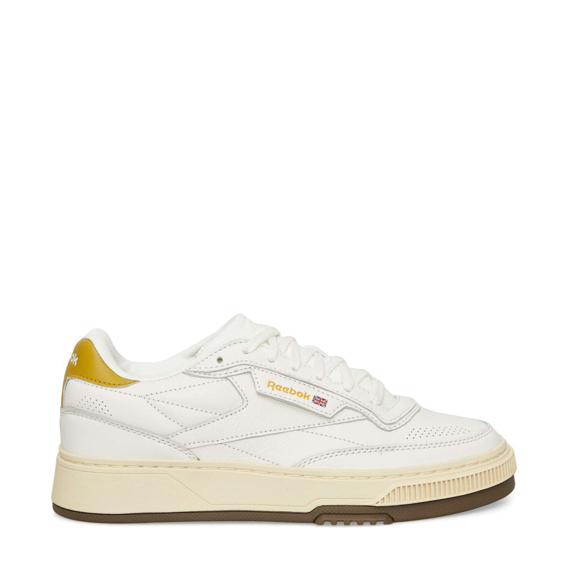 A white Reebok sneaker with a cream-colored sole, brown outsole, and gold accents on the heel and logo. The shoe features a low-top design and visible stitching details.