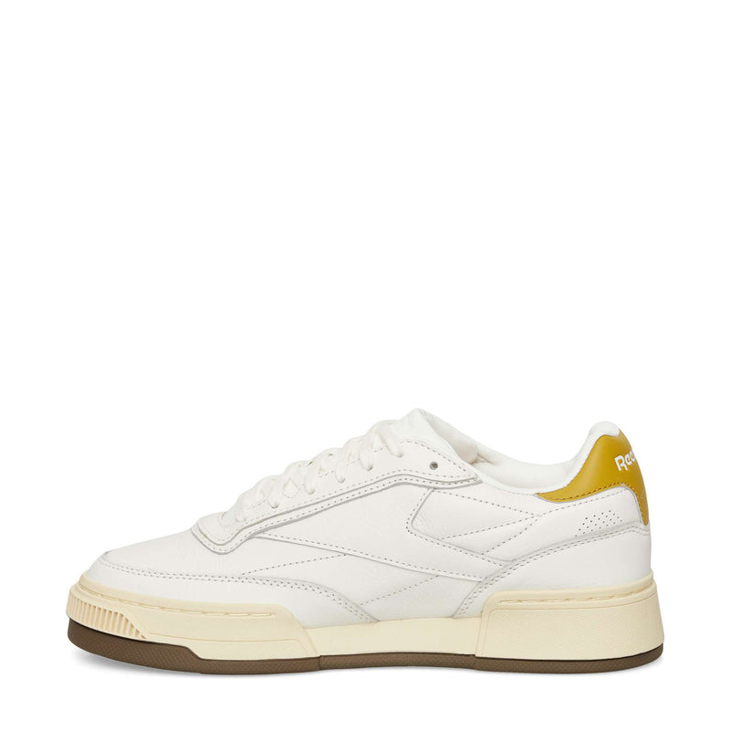A white low-top sneaker with off-white and gum-colored sole, subtle stitching details, and a mustard yellow accent on the heel featuring the Reebok logo.