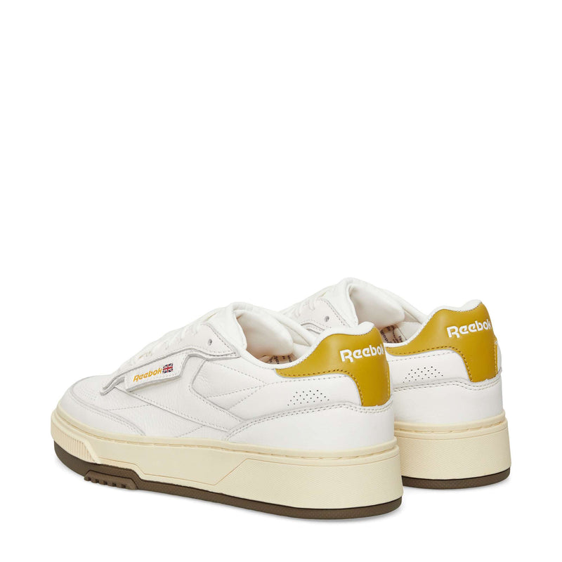A pair of white Reebok sneakers with yellow heel accents, beige soles, and brown outsoles, shown from the back and side on a plain white background.