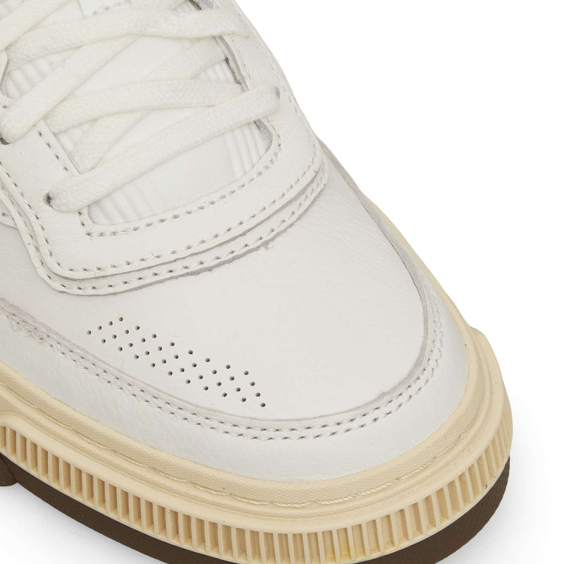 Close-up of a white sneaker with white laces, perforated detailing on the toe area, and an off-white textured midsole with a brown outsole.