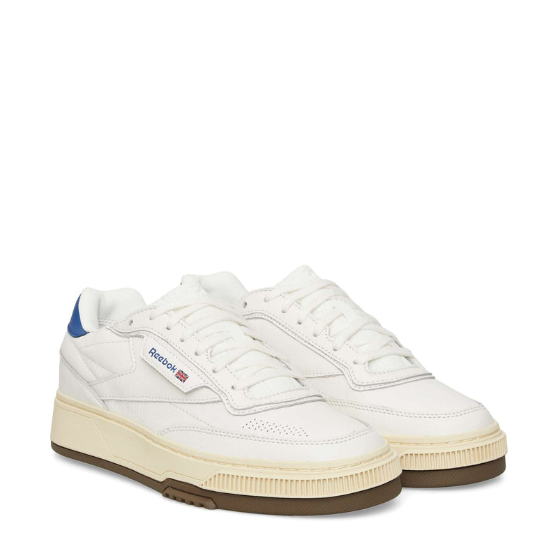 A pair of Reebok Club C LTD Shoes in white with blue heel accents, a beige midsole, and brown outsole is displayed on a white background. The Reebok logo and British flag detail complete this classic lifestyle tennis shoe.