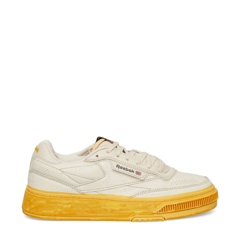 A left-side view of the Reebok Club C LTD Shoes in beige, featuring a yellow sole, the iconic Reebok logo, and a small British flag on the side—an updated take on the classic Club C tennis shoe.