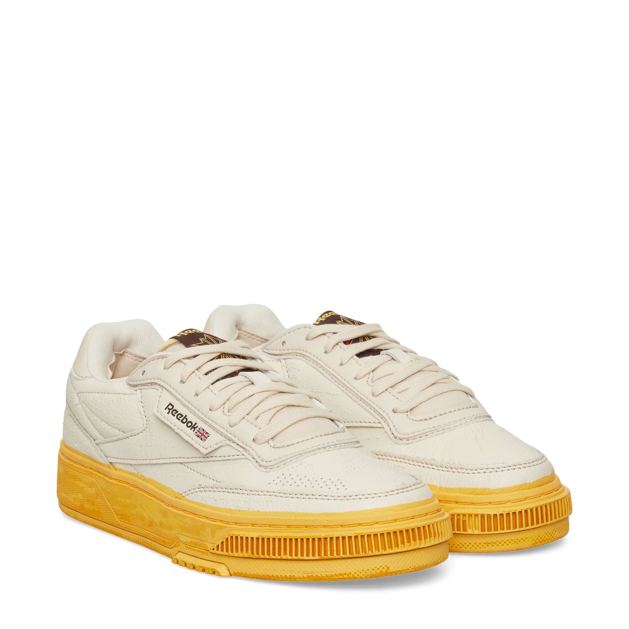 The Reebok Club C LTD Shoes are cream-colored with yellow soles, white laces, and a retro-inspired design. They feature the Reebok logo and Union Jack on the side, blending classic tennis shoe style with modern flair.