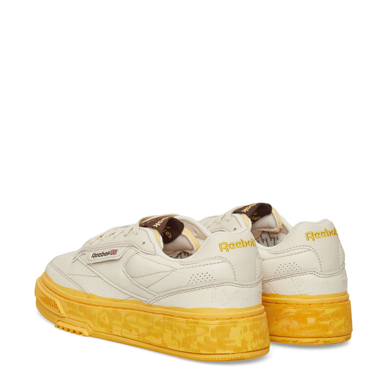 The Reebok Club C LTD Shoes feature a cream upper, yellow patterned soles, brown tongue labels, a yellow Reebok logo on the back, and black and red details on the side for a classic lifestyle look.