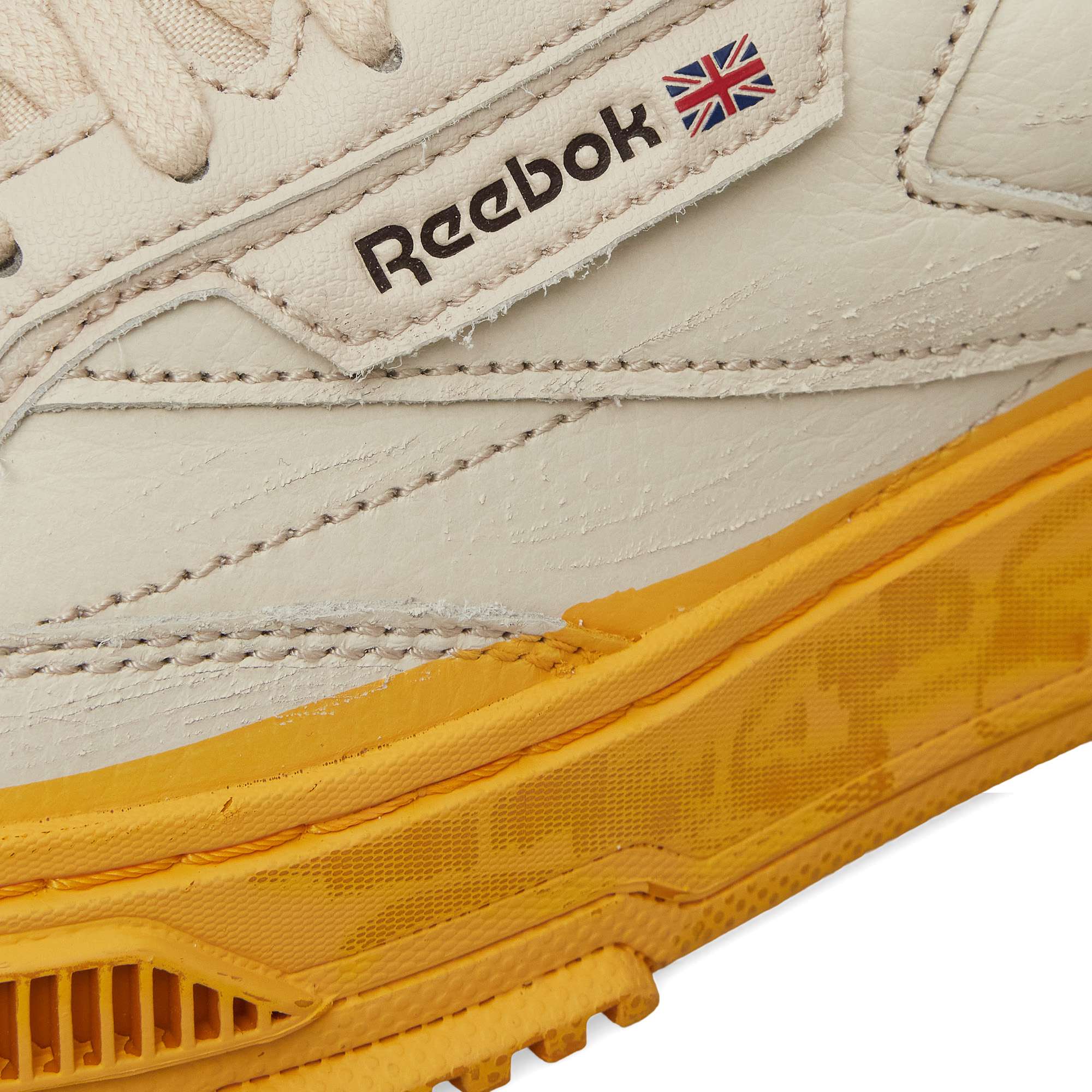 Close-up of the Reebok Club C LTD Shoes in white, showcasing the Reebok logo, UK flag, and a textured yellow rubber sole. Classic stitching details accent the leather upper for this timeless lifestyle sneaker.