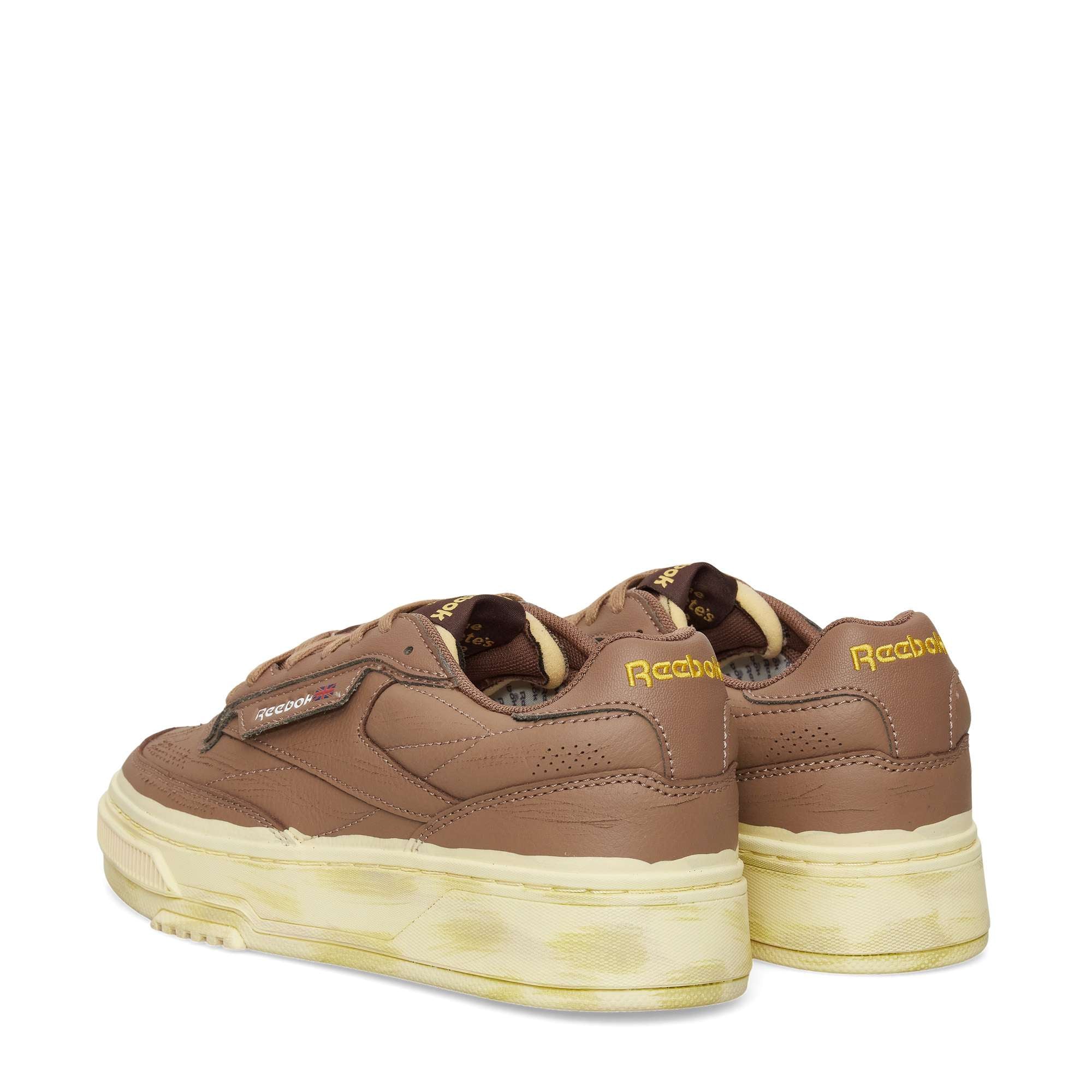 A pair of Reebok Club C LTD Shoes in brown with yellow logo accents and thick off-white soles, featuring a vintage-inspired, slightly worn look—shown from both back and side angles.