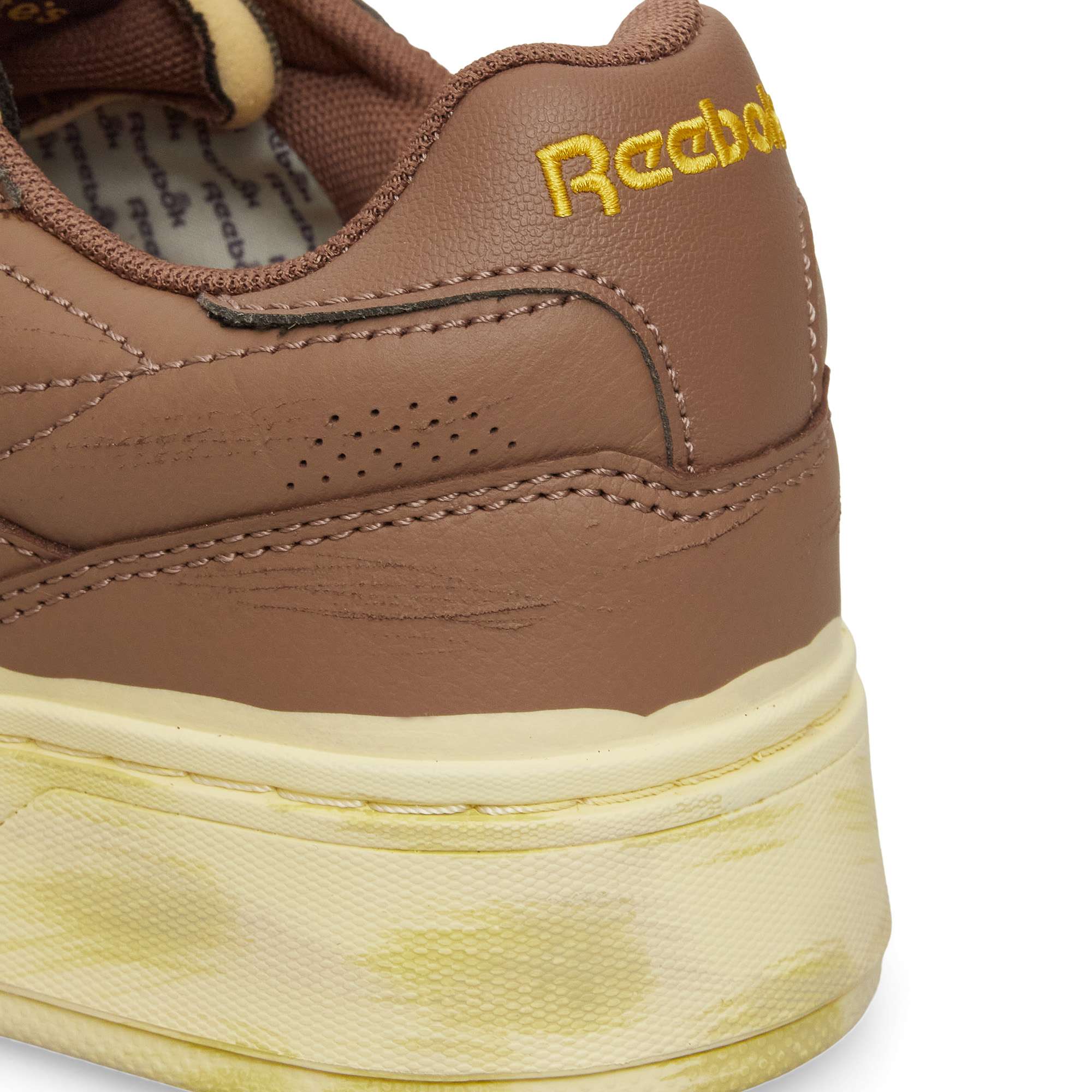 Close-up of the back of a brown Reebok Club C LTD shoe with yellow embroidered branding and a cream-colored midsole displaying yellowish stains, highlighting the timeless appeal of this classic from Reebok.