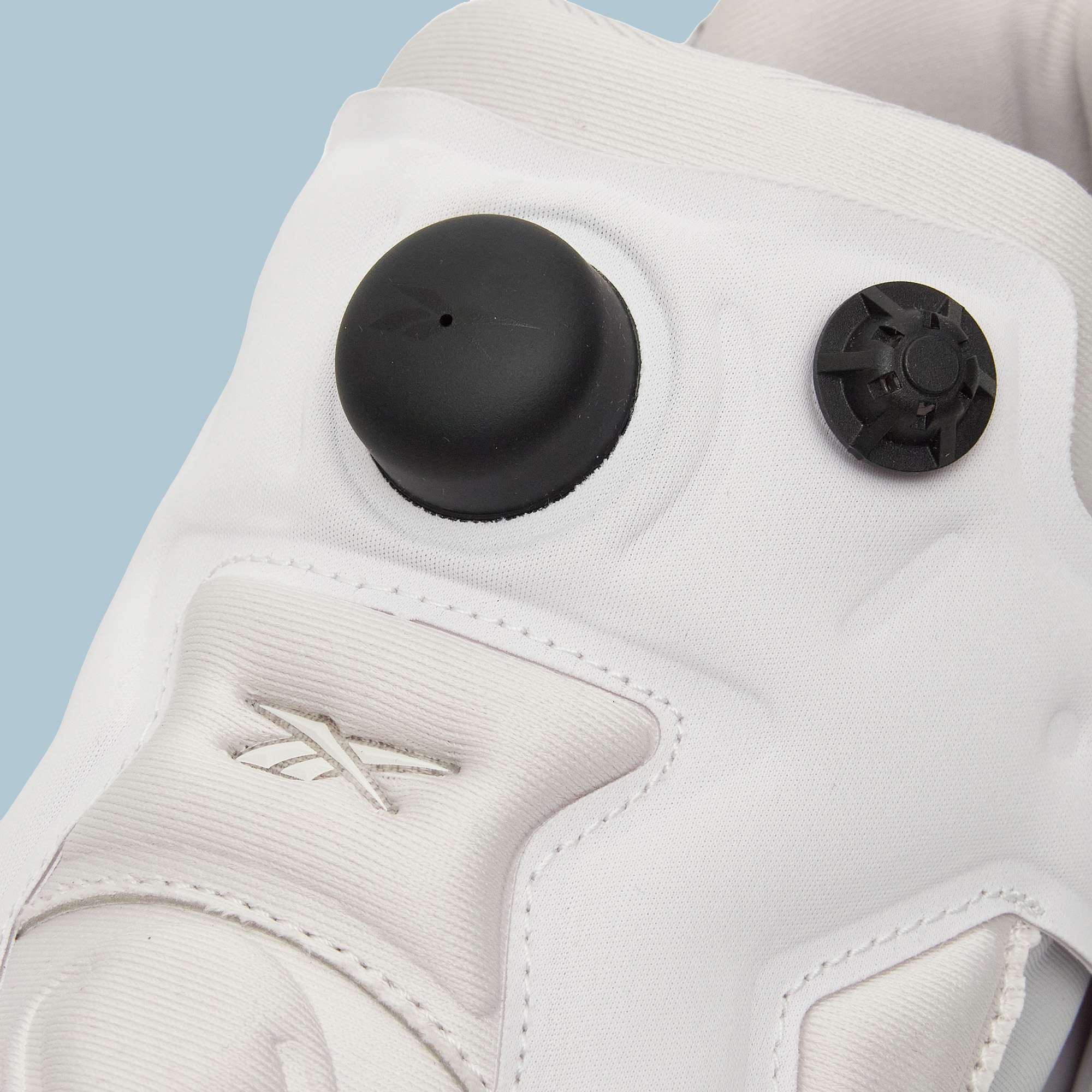 Close-up of a white Reebok Instapump Sliver Shoe with a black circular air pump and smaller black valve on the tongue. The Reebok logo is stitched in white on the side, set against a light blue background.