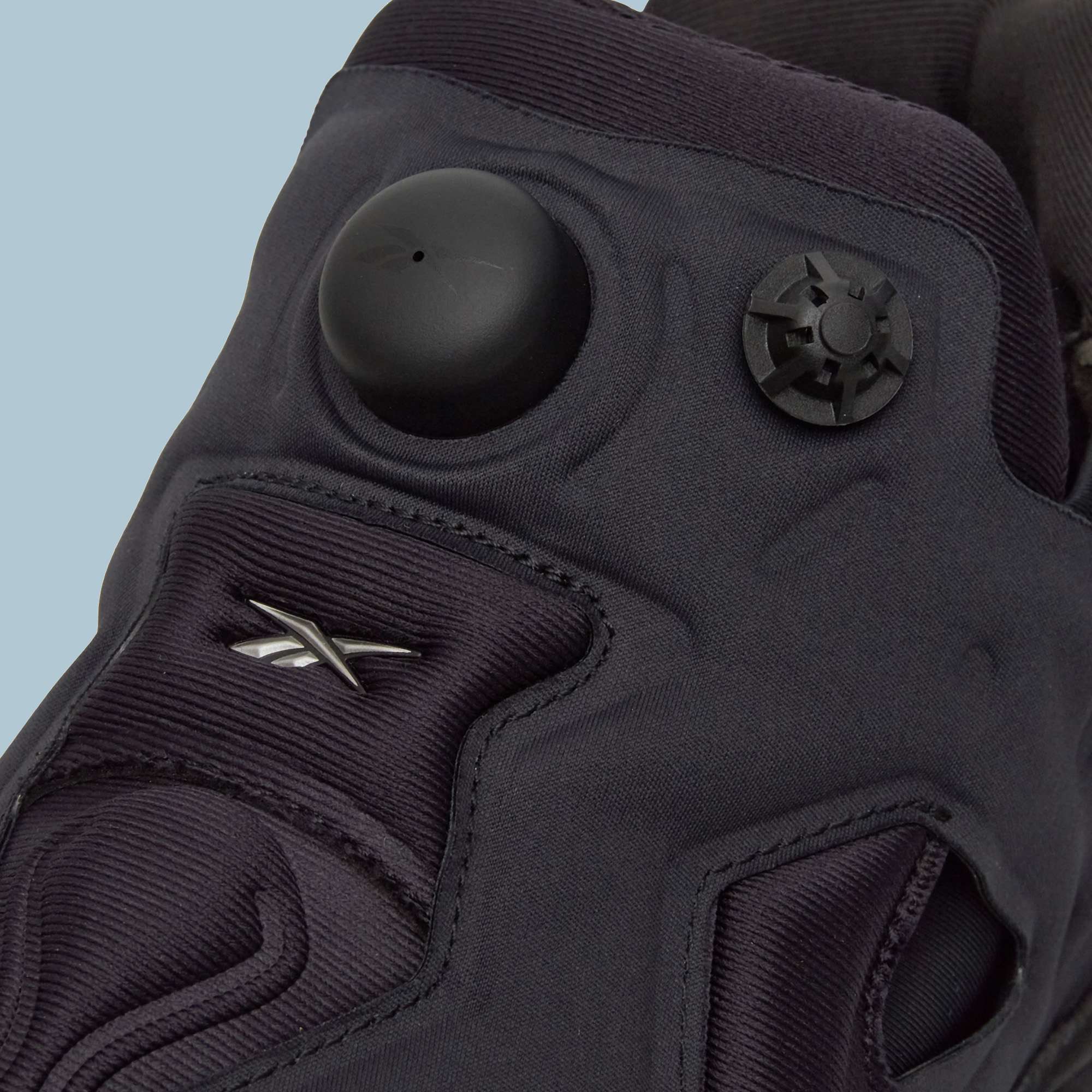 Close-up of a black Reebok Instapump Sliver Shoe featuring the air pump system, rounded button, star-shaped release valve on the tongue, and signature logo against a light blue background.