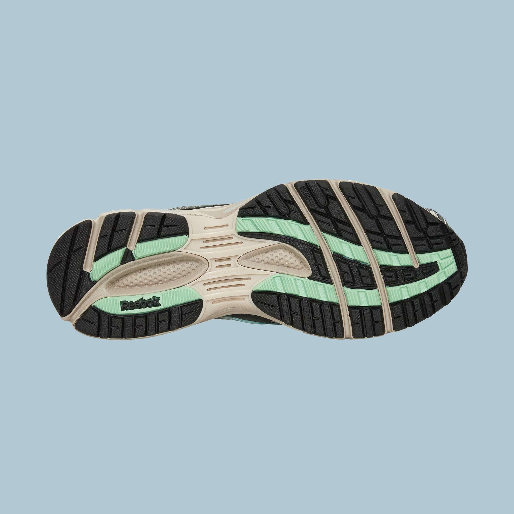 The image displays the sole of Reebok’s Premier Road Ultra Shoes, featuring black, beige, and mint green accents, multiple grooves, a dual-density midsole, textured patterns, and the Reebok logo on the heel against a light blue background.