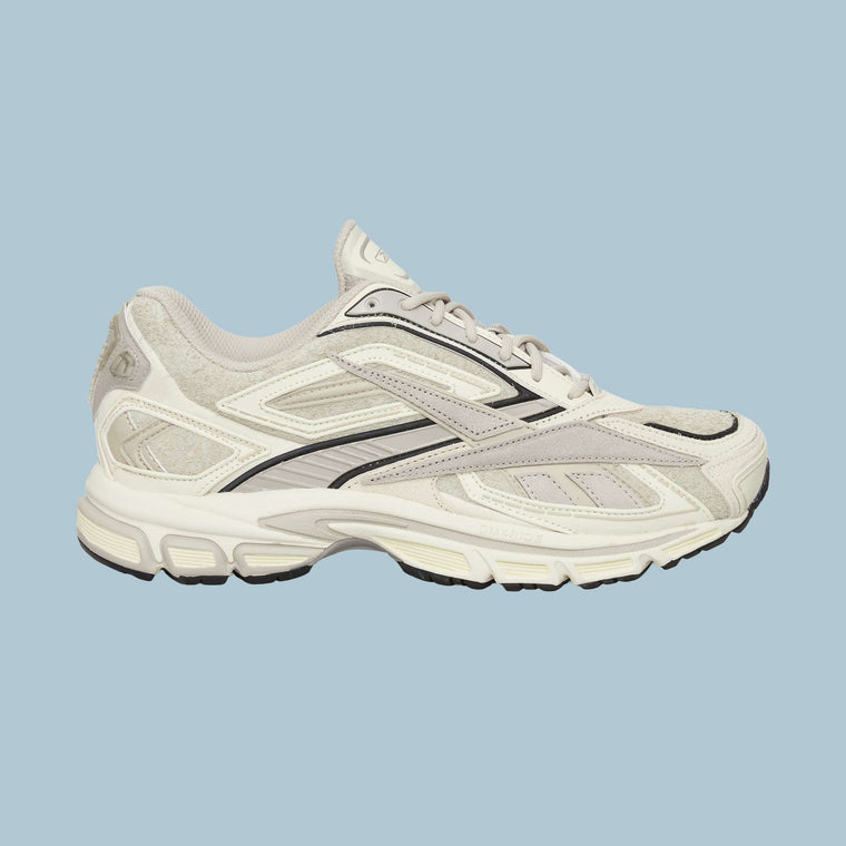 The Reebok Premier Road Ultra Shoes feature beige and white uppers with black accents, mesh panels, and a textured sole with a dual-density midsole, shown against a light blue background.