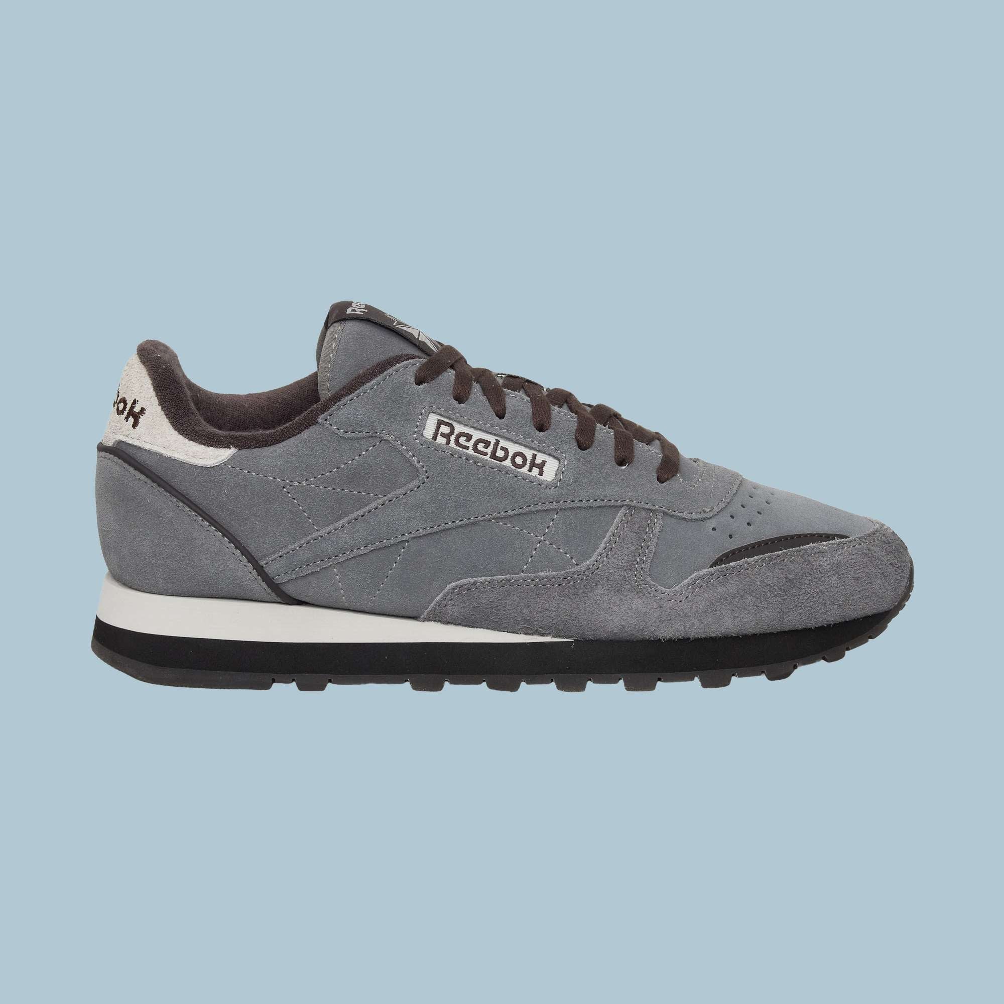 The Reebok Classic Leather Premium LTD Shoes feature a gray upper, black laces, a white and black sole, and a white heel accent—showcasing their retro-inspired silhouette against a light blue backdrop.