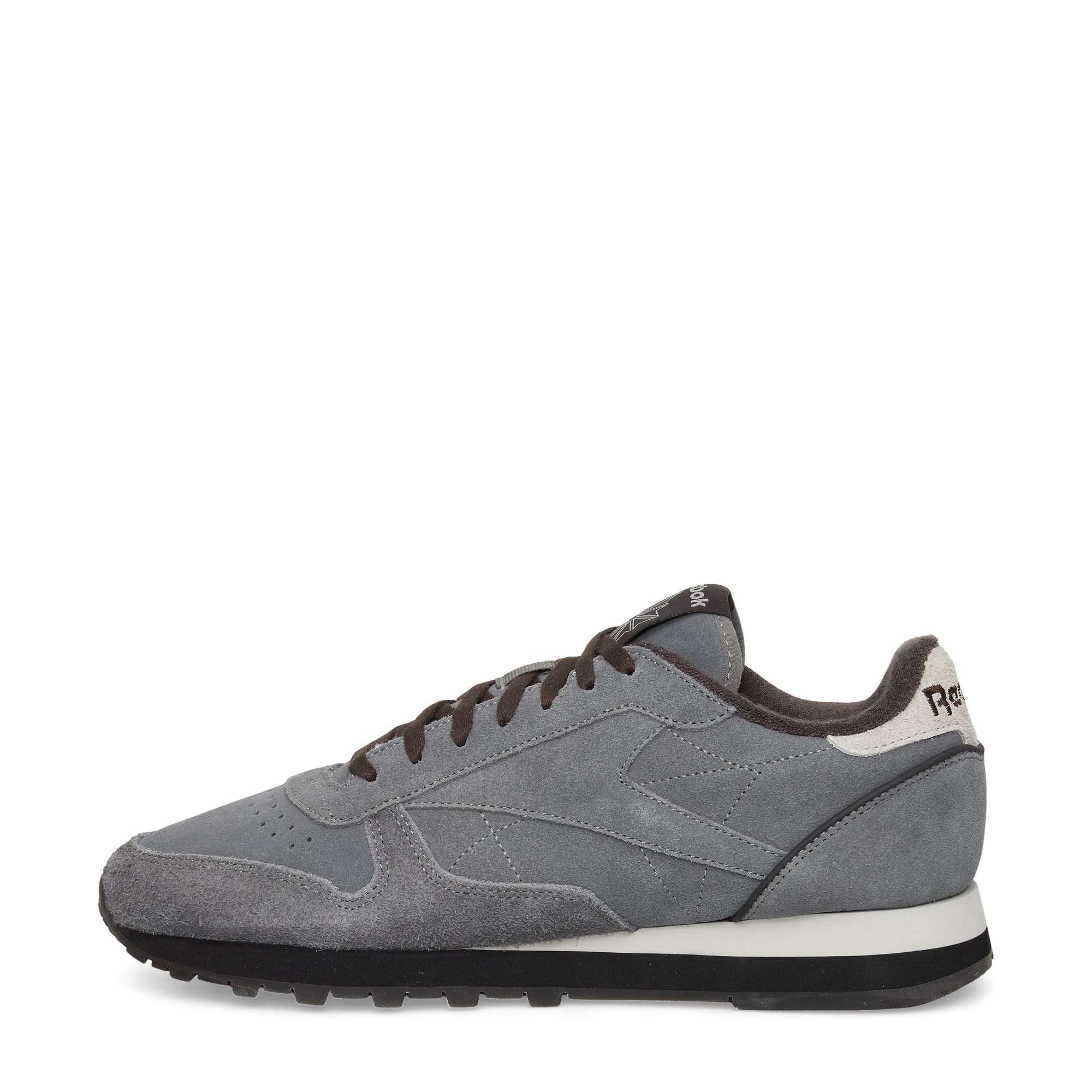 A side view of a gray suede sneaker with black laces, a white midsole, black outsole, and black interior lining. The shoe has subtle stitching details and a small logo on the heel.
