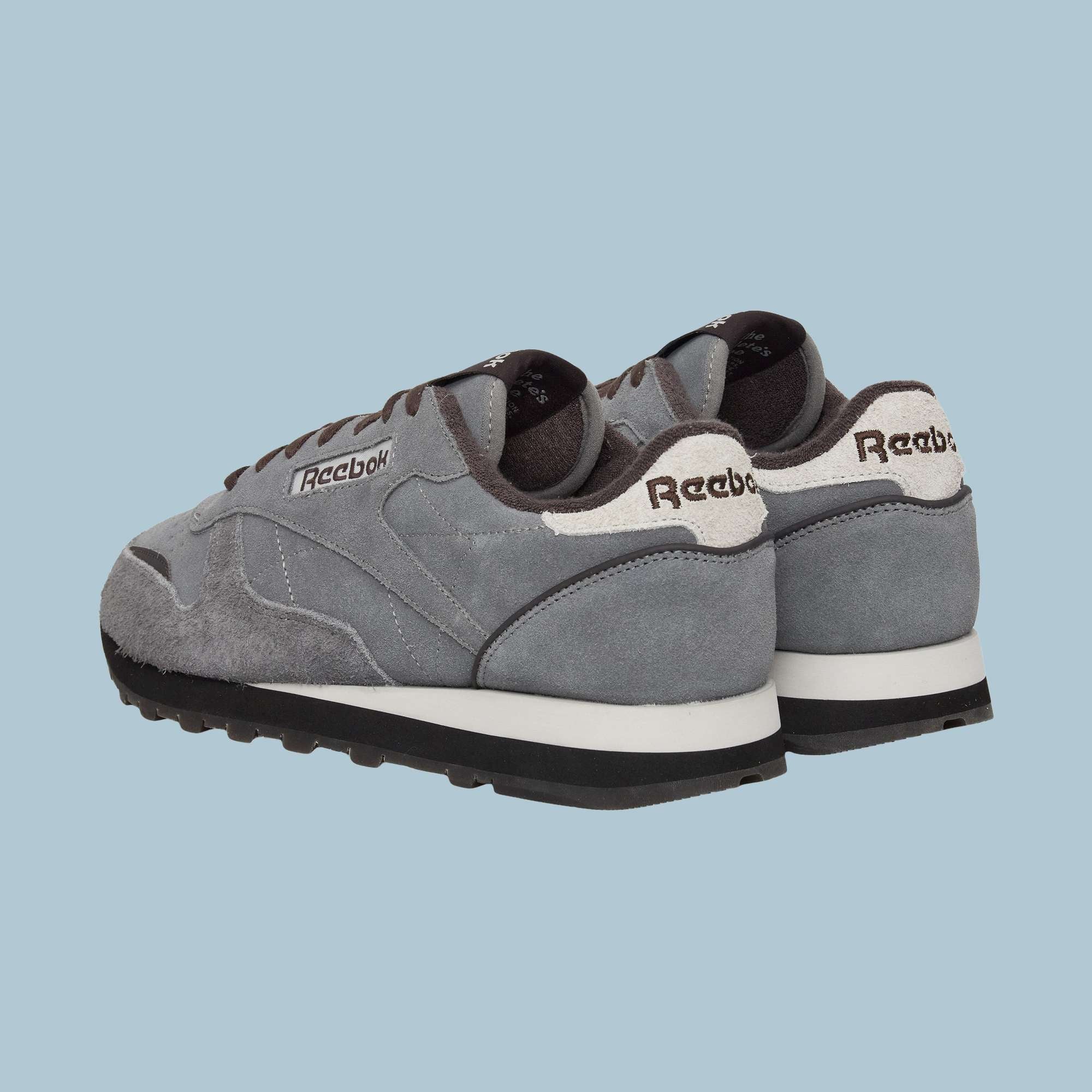 A pair of Reebok Classic Leather Premium LTD Shoes in gray, featuring white soles and black bottoms, are displayed on a light blue background with back and side views that highlight their retro, timeless silhouette.