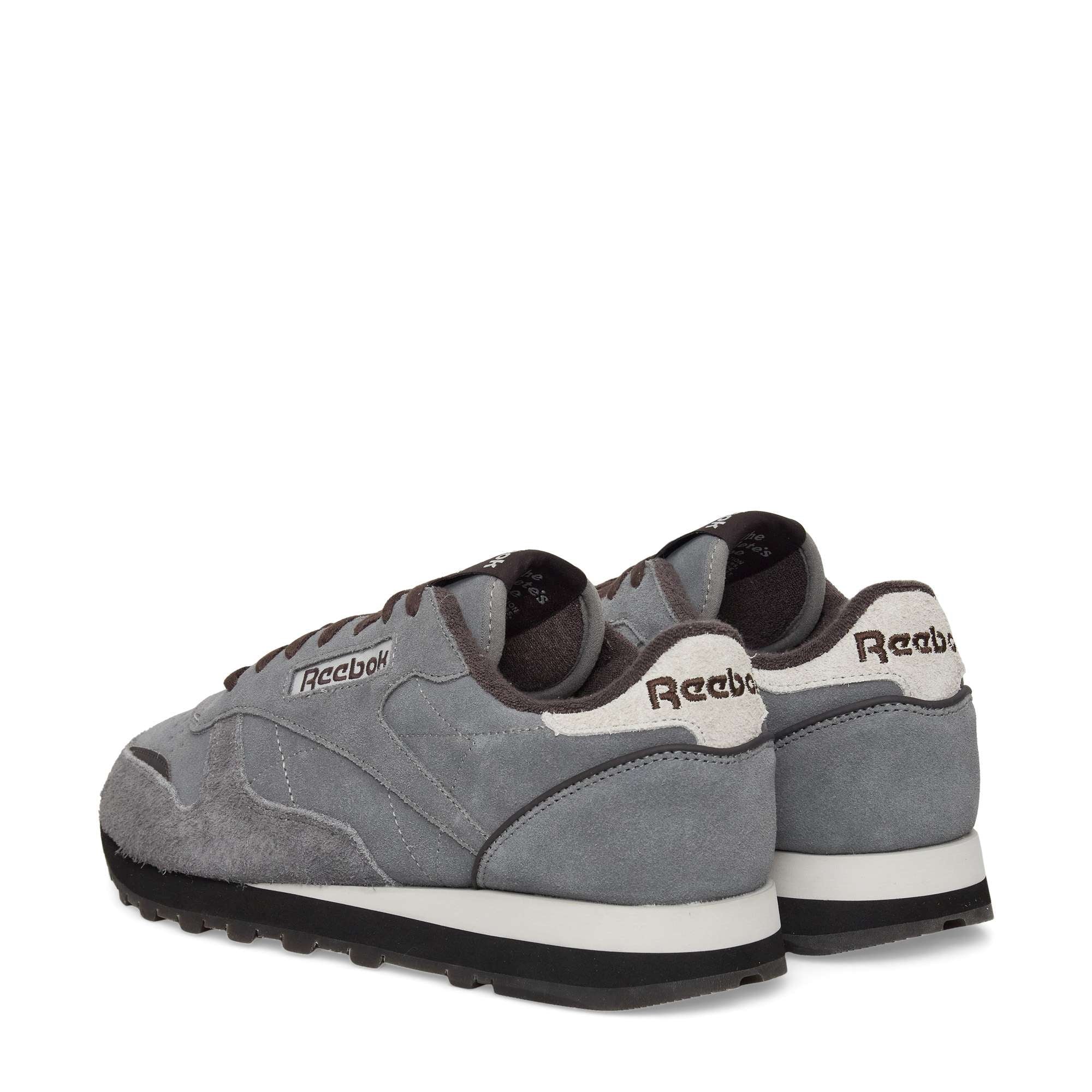 A pair of gray Reebok sneakers with white and black soles, featuring white accents and Reebok branding on the heel. The sneakers are positioned angled to display the back and side view.