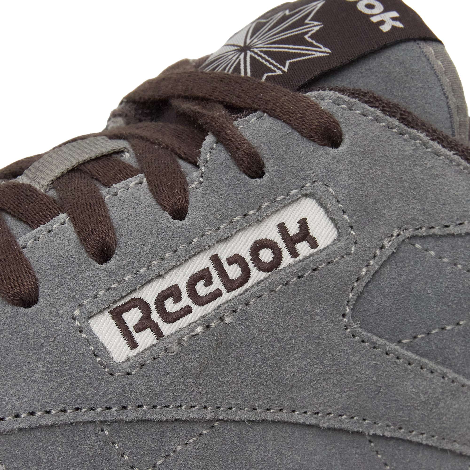 Close-up of a gray suede Reebok sneaker with brown laces, showing the stitched Reebok logo and part of a maple leaf design on the tongue.