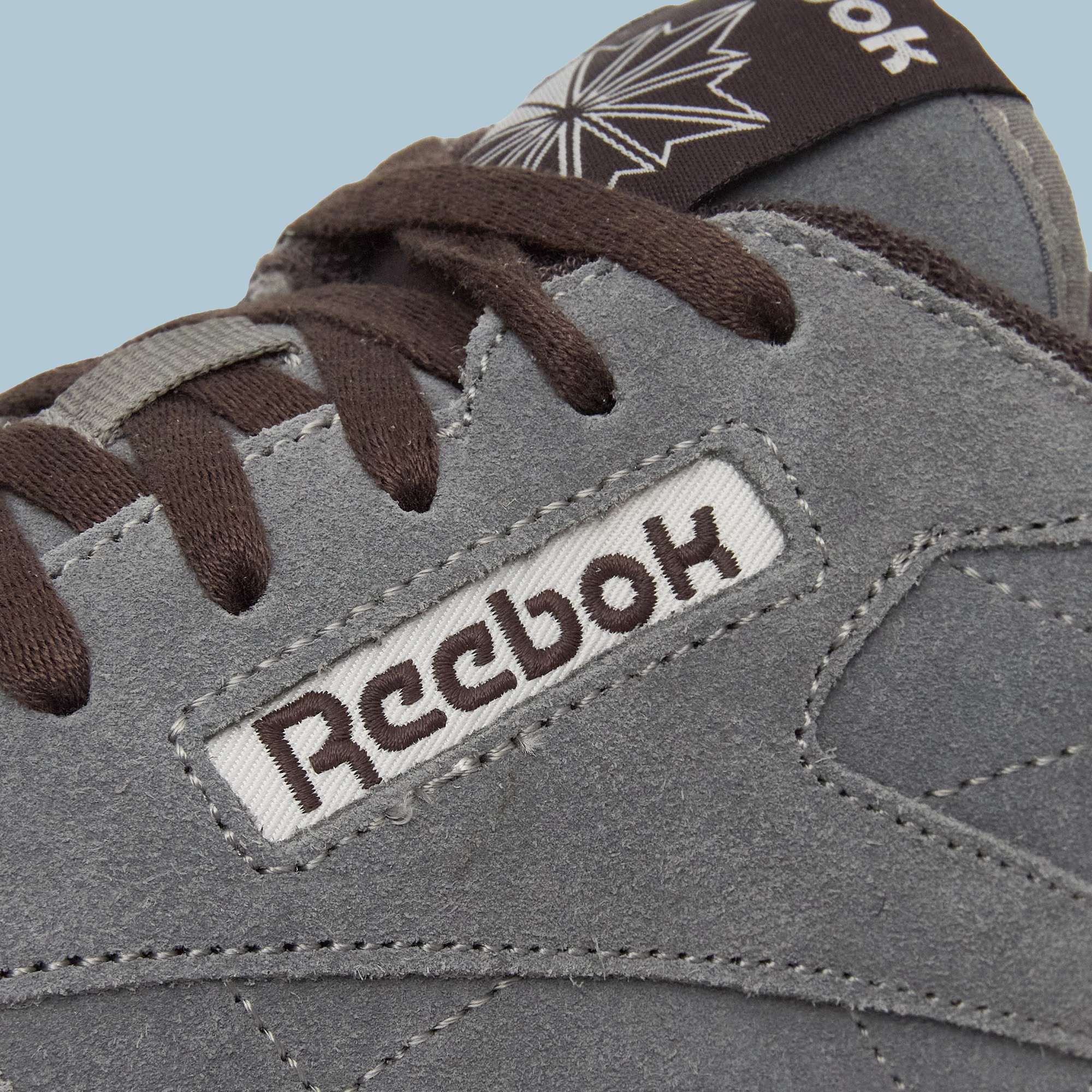 Close-up of a grey suede Reebok Classic Leather Premium LTD Shoe with brown laces. The Reebok logo and part of a star logo are visible, highlighting its classic silhouette against a light blue background.