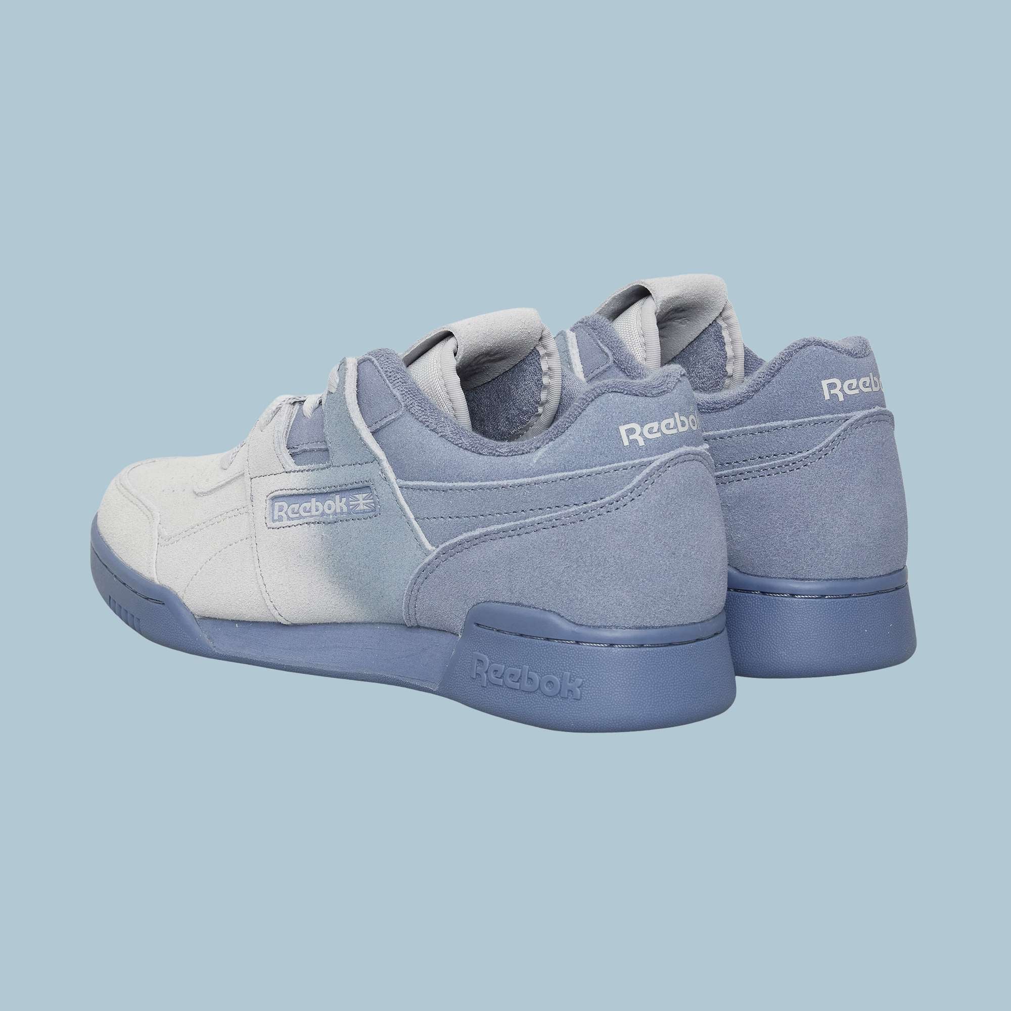 A pair of Reebok Workout Plus OG Shoes in light blue and gray are displayed on a light blue background, showing back and side views with visible Reebok logos that highlight the classic silhouette.
