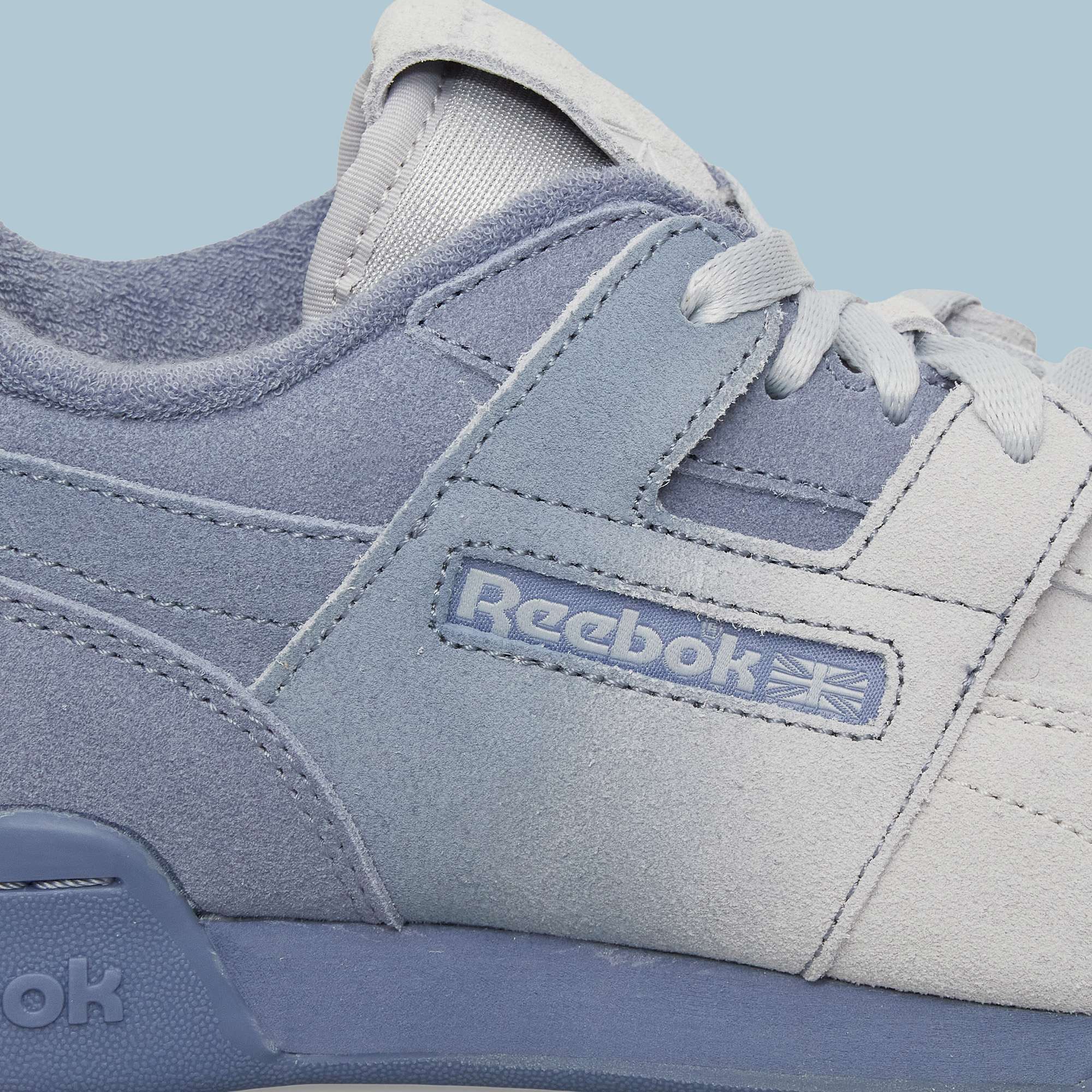 Close-up of light blue and white Reebok Workout Plus Og Shoes with visible side logo, laces, and stitching. The classic retro silhouette stands out against a light blue background.