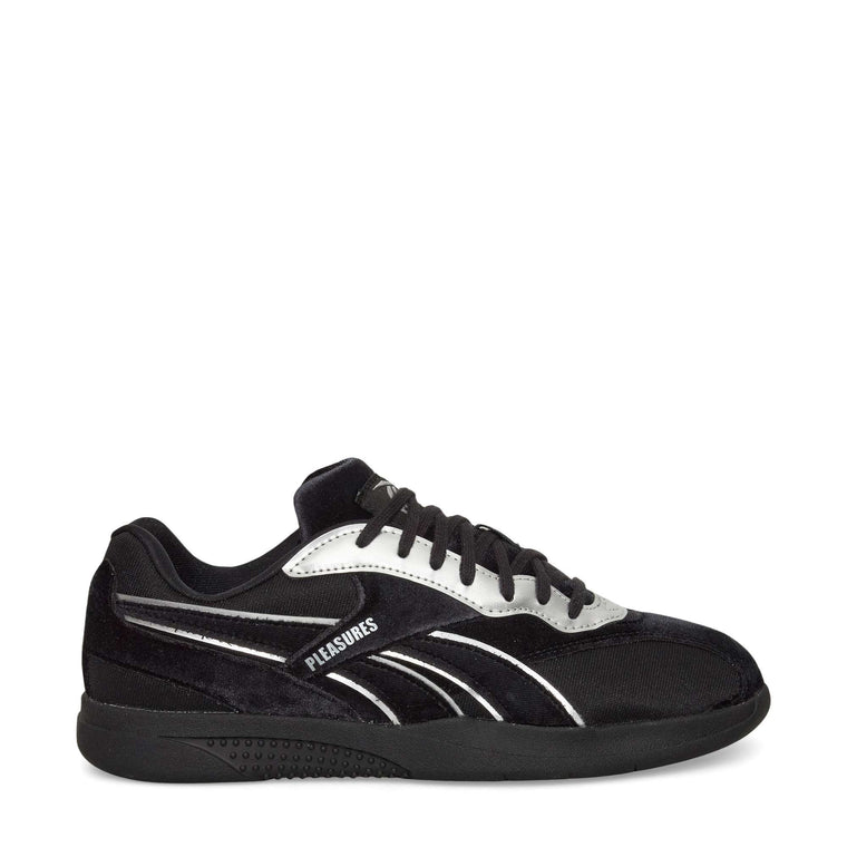 A black low-top sneaker with black laces, silver accents, and the word PLEASURES printed on the side. The shoe has a black sole and a sleek, modern design.