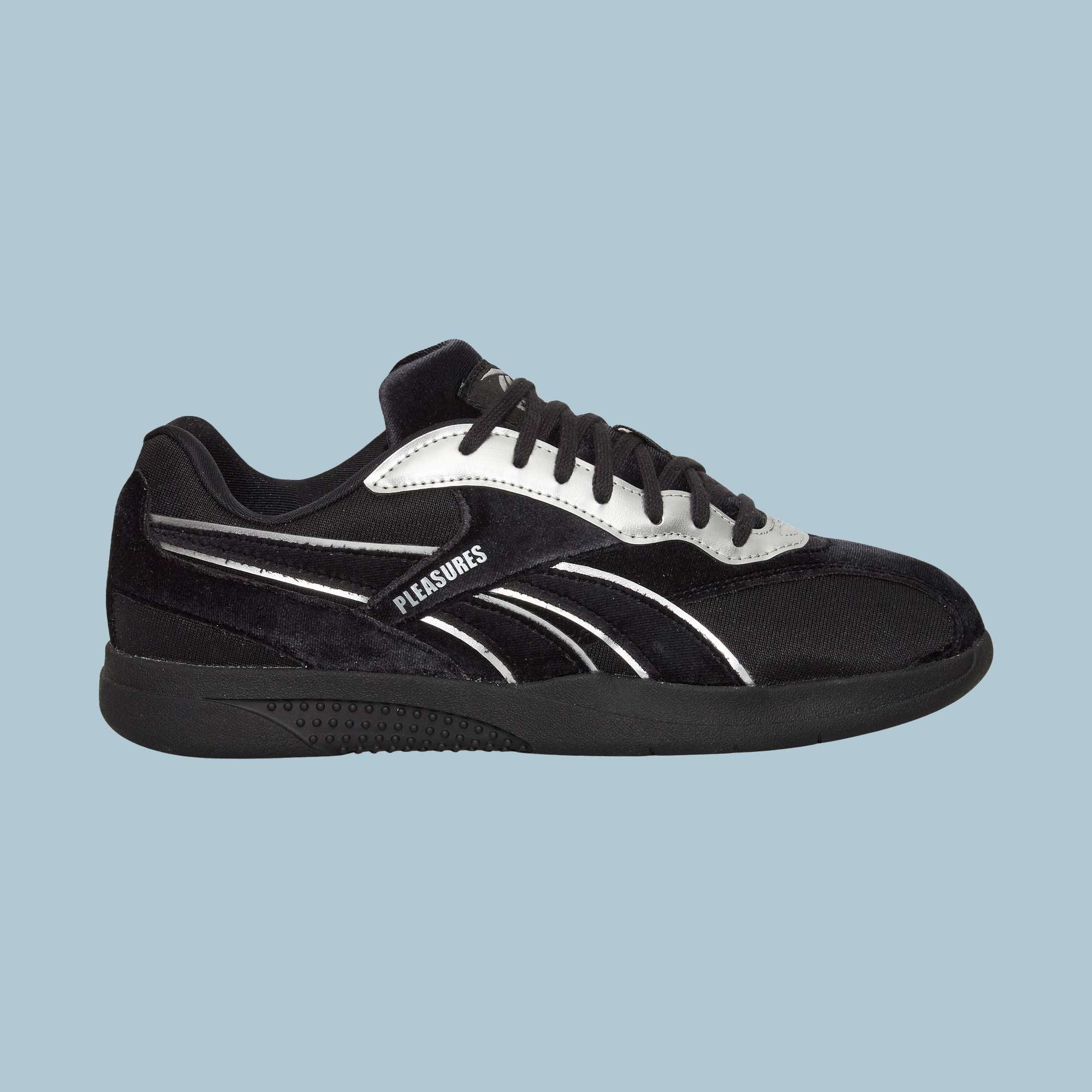 The Reebok Pleasures Hammer Street Shoes, a black sneaker with a velvet upper and white and silver accents, features "PLEASURES" on the side—part of the PLEASURES collaboration—set against a plain light blue background.