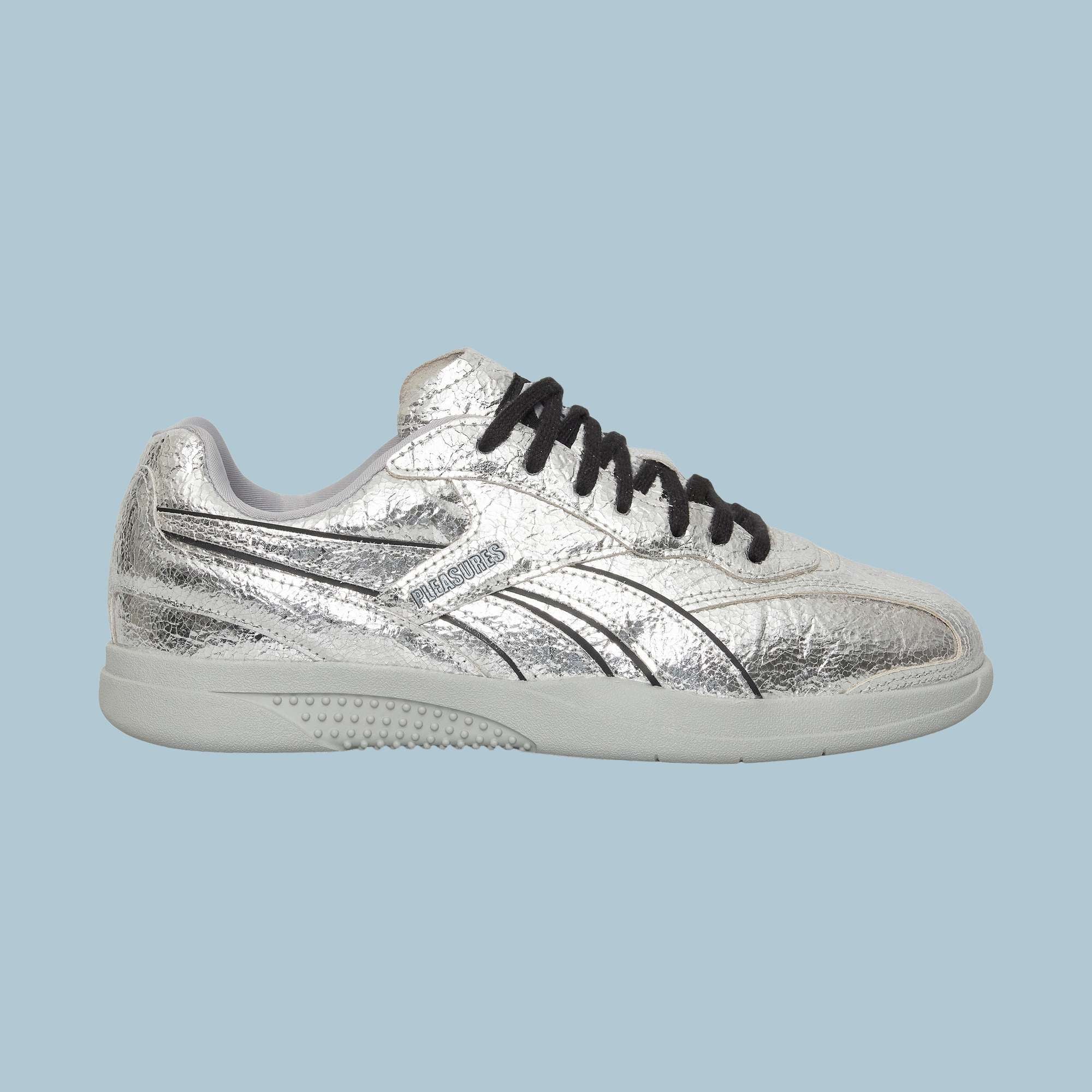 The Reebok Pleasures Hammer Street Shoes, shown in silver leather with black laces, a white sole, black line accents, and a subtle textured finish, are inspired by classic Reebok indoor soccer shoes against a light blue background.