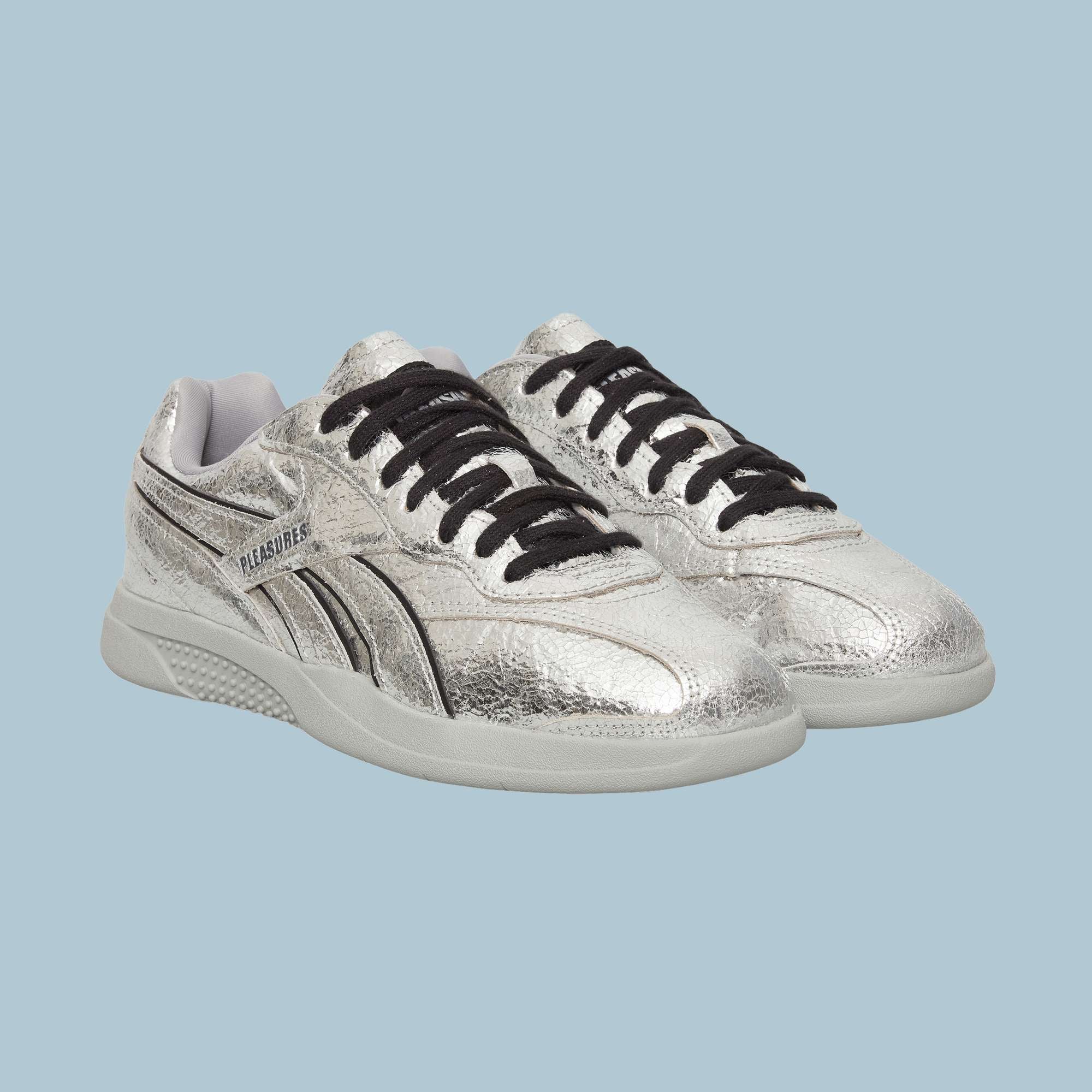 The Pleasures Hammer Street Shoes by Reebok are silver leather sneakers with black laces and white soles, featuring a textured finish and side logo detail, inspired by classic Reebok indoor soccer styles. Displayed on a light blue background.