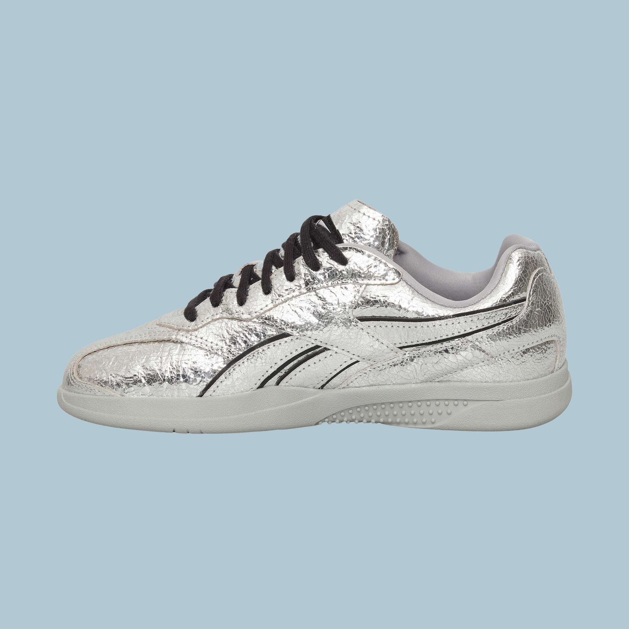 The Reebok Pleasures Hammer Street Shoes, featuring shiny silver leather, black laces, and swooping design details, are displayed from the side on a light blue background.