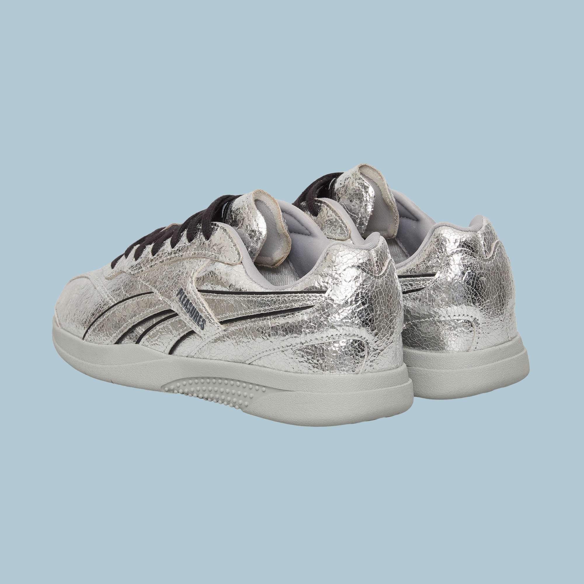 A pair of Reebok Pleasures Hammer Street Shoes in metallic silver leather with black stripes and accents, shown from the back angled left, on a light blue background—part of a PLEASURES collaboration.