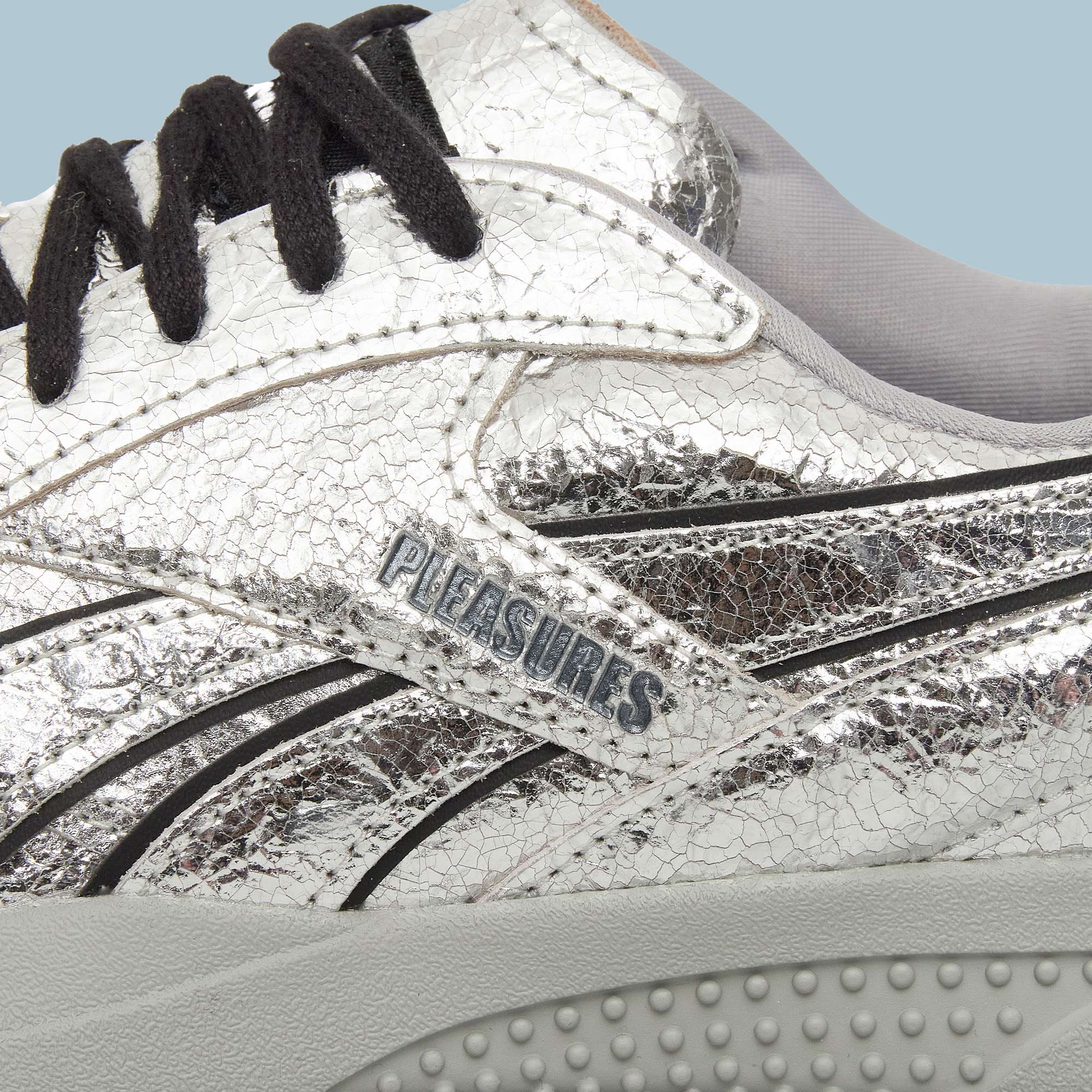 Close-up of Reebok Pleasures Hammer Street Shoes in silver leather with a cracked texture, black laces, light gray inner lining, and Pleasures branding on the side above a textured light gray sole.