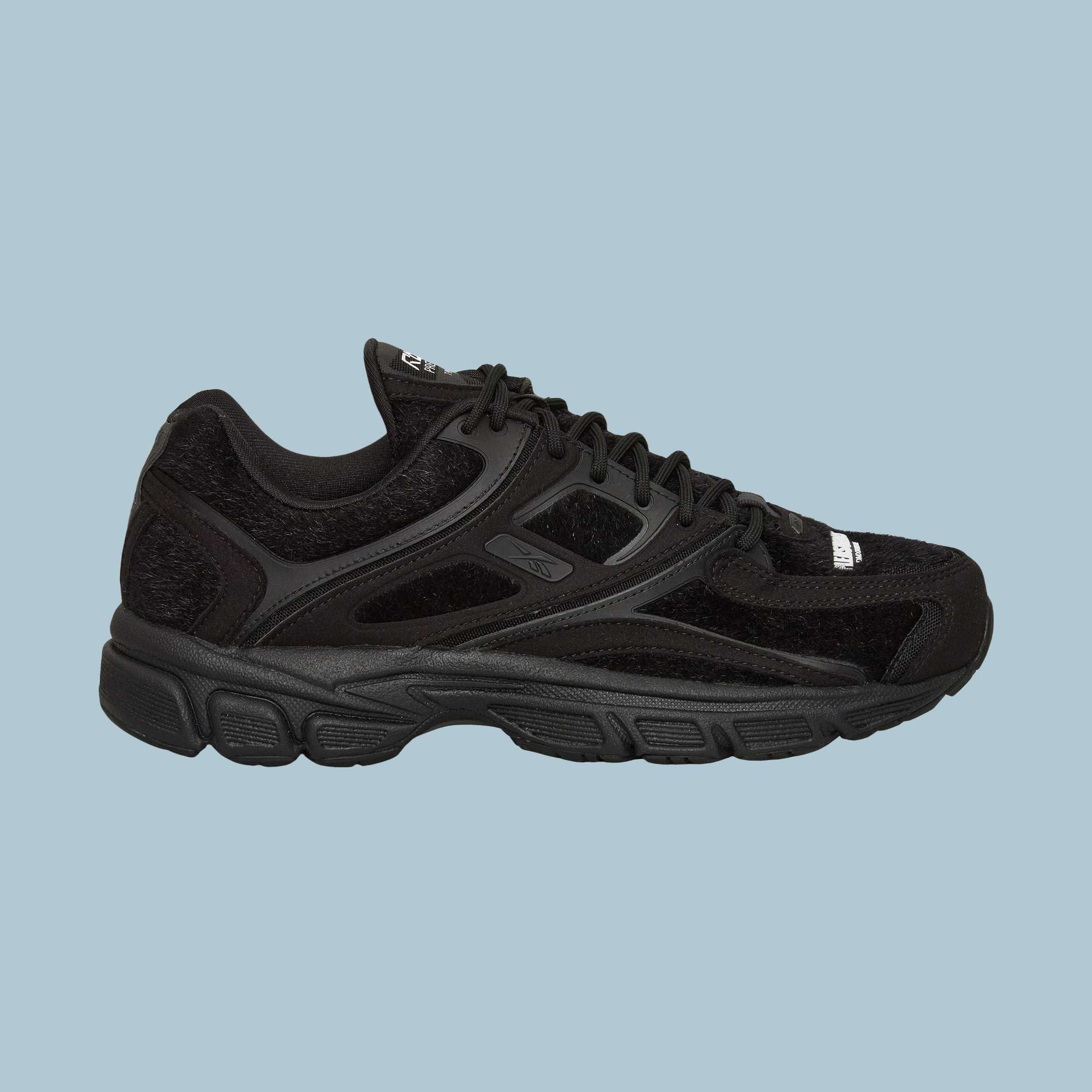The Reebok Pleasures Premier Trinity KFS Shoes feature a black athletic design with mesh and suede details, black laces, a textured rubber sole, and subtle Reebok branding. Displayed on a light blue background.