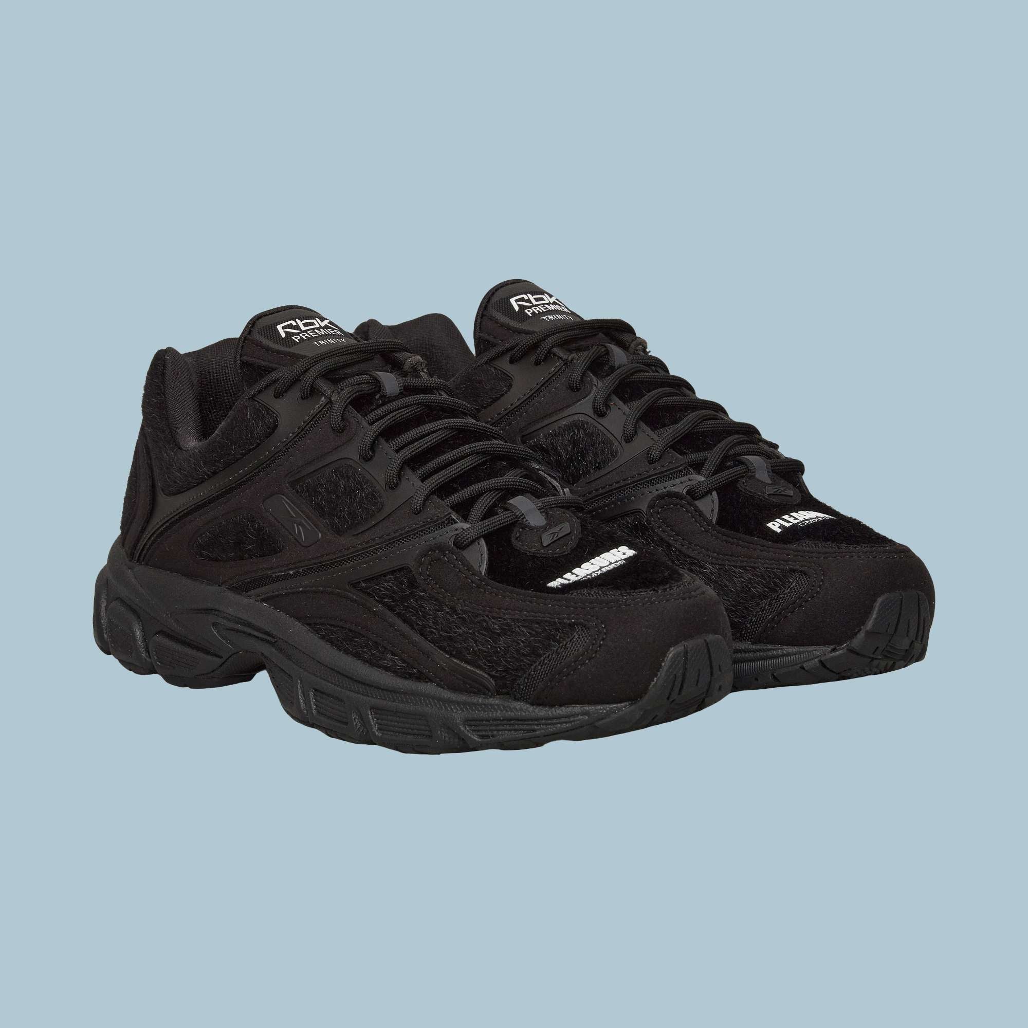 The Reebok Pleasures Premier Trinity KFS Shoes are black athletic sneakers with thick soles, mesh accents, and a unique pony hair upper, displayed on a plain light blue background.