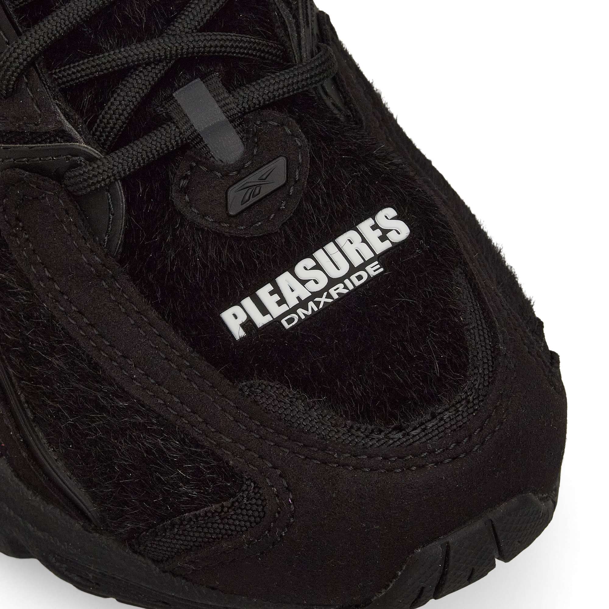 Close-up of a black sneaker with textured details. The words PLEASURES DMXRIDE are printed in white on the upper part of the shoe near the laces, with a small Reebok logo above.