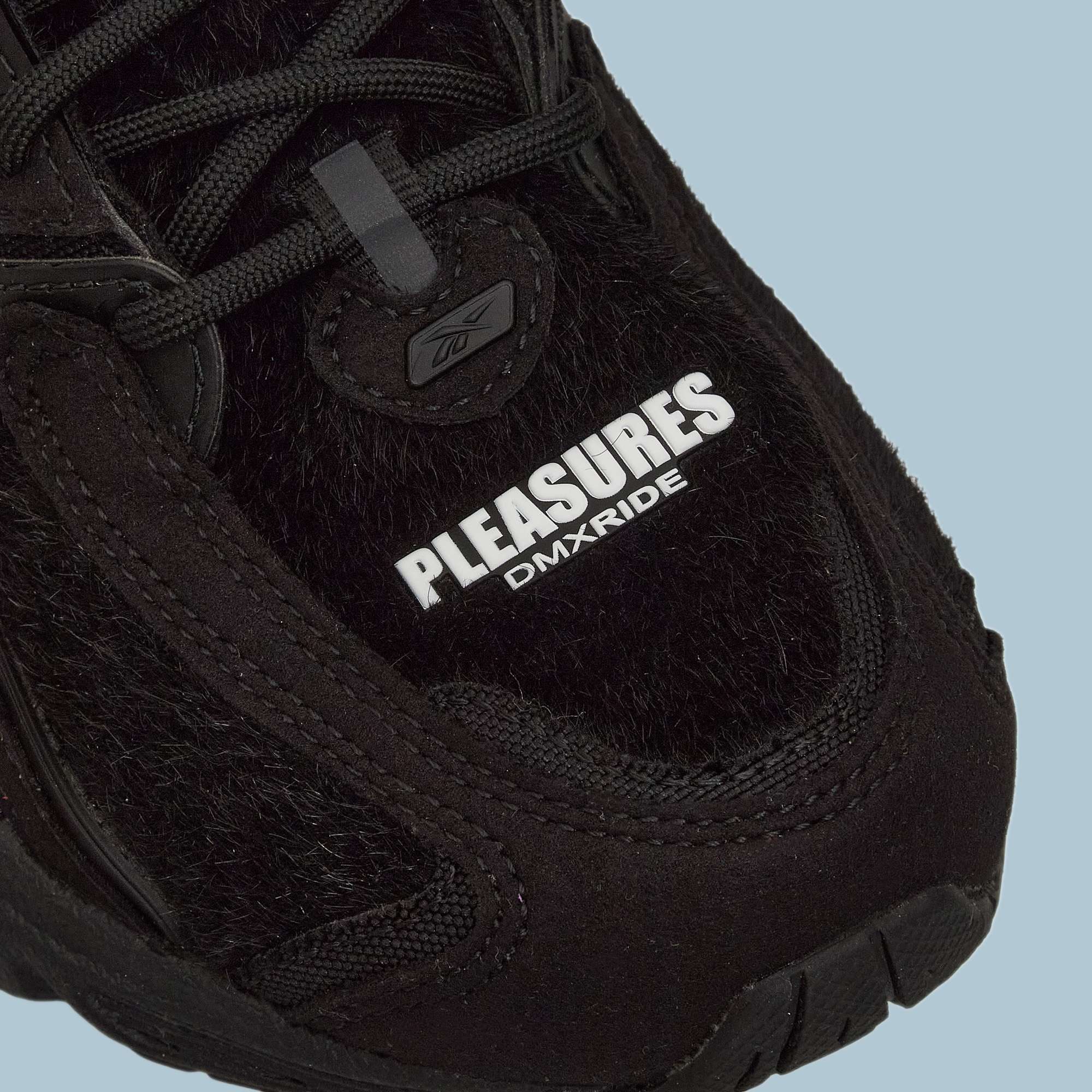Close-up of the Pleasures Premier Trinity KFS Shoes from Reebok, featuring a black textured pony hair upper. "PLEASURES" stands out in bold white with DMXRIDE underneath. Finished with black laces and stitching for a striking look.