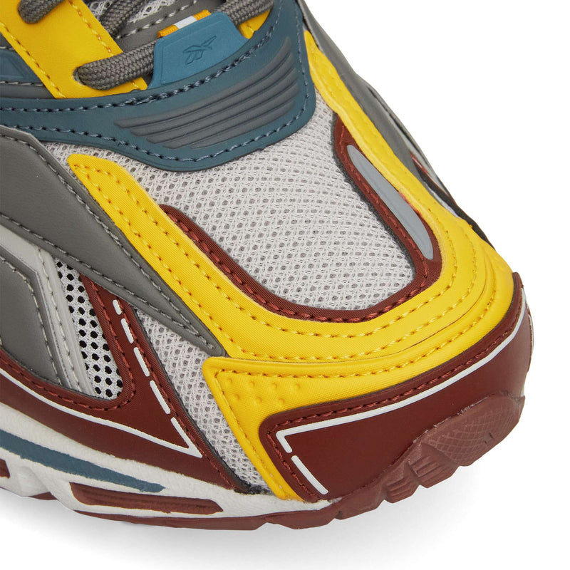 Close-up of the Reebok Premier Road Ultra shows mesh fabric, yellow, grey, blue, and brown overlays, visible stitching, and a textured rubber sole with dual-density midsole.