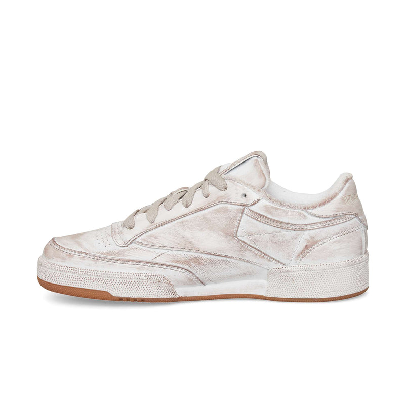 A side view of the Reebok Club C 85 Vintage, a low-top sneaker with a worn white finish, gray laces, textured leather details, and a brown rubber sole. The distressed look adds timeless appeal to this classic tennis-inspired design.