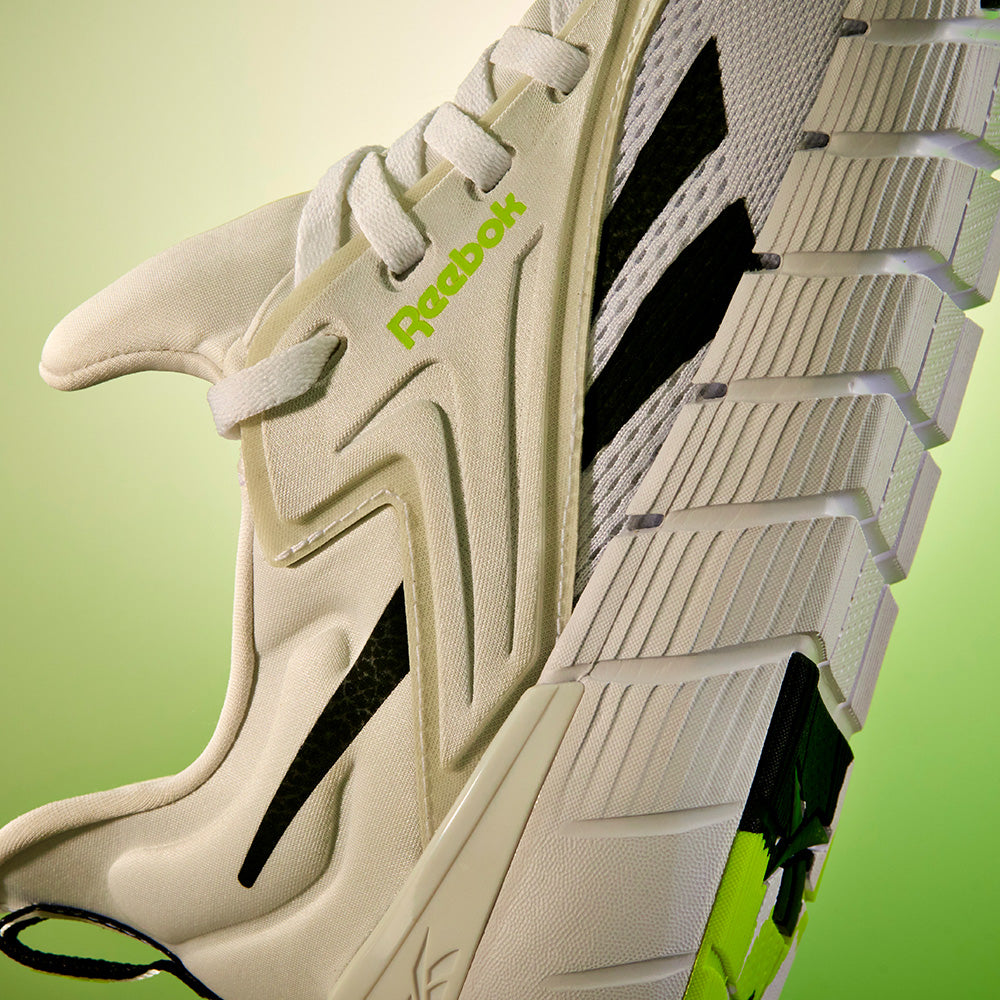 Close-up of Reebok Women's Nano Gym Training Shoes in black with neon green accents, detailed tread, and brand logo—ideal for fitness classes—displayed against a light green gradient background.