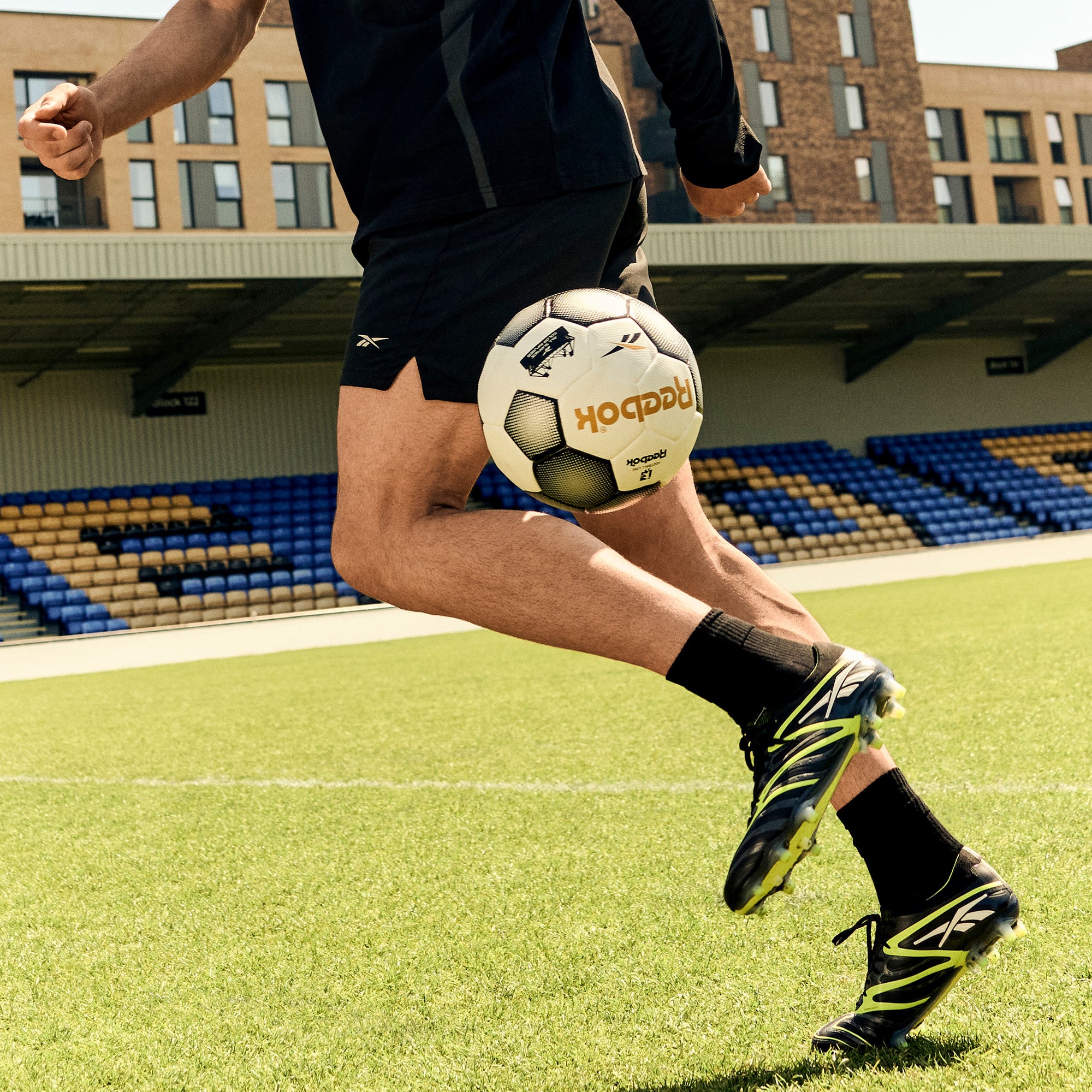 Wearing Reebok Sidewinder 26 Elite FG Cleats, a soccer player in black gear juggles a black and white ball on firm grass before empty stadium seats.