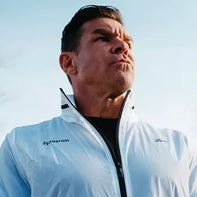 A man with short dark hair wearing a white jacket looks off into the distance with a serious expression against a blue sky background.