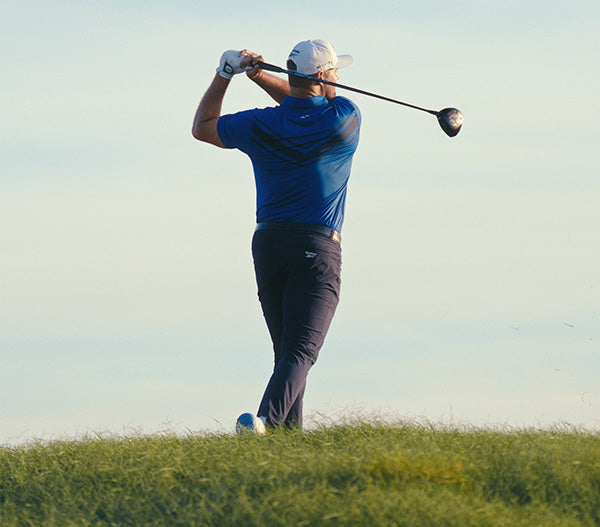 A golfer wearing a blue shirt and navy pants stands on a grassy hill, finishing a swing with a driver club as he faces away from the camera under a clear sky.