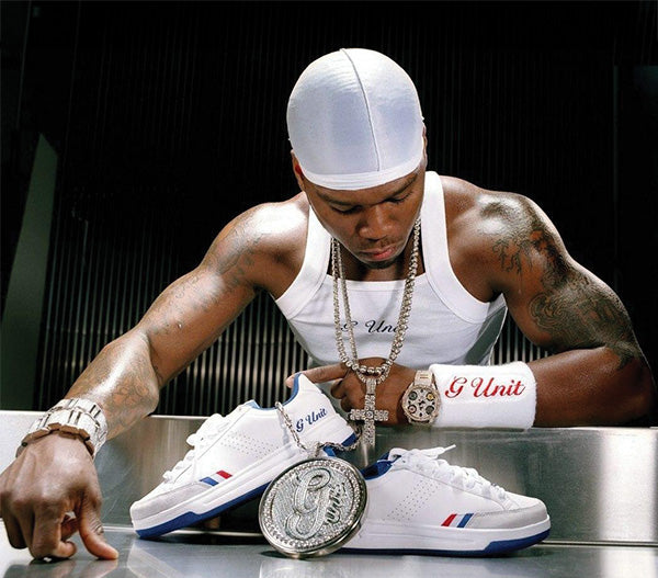 A man wearing a white cap, white tank top, and multiple gold chains leans over a metallic surface while looking at a pair of white G-Unit sneakers with blue and red accents. A large silver coin rests in front of the shoes.