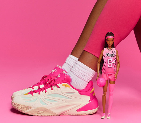 A pair of Reebok athletic sneakers in pink, white, and pale blue, shown worn on a foot with white socks, alongside a Barbie basketball doll in a matching pink outfit against a solid pink background.