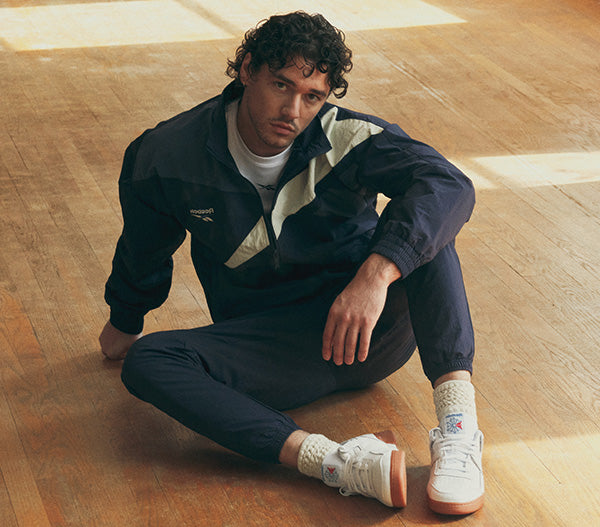 A man sits on a wooden floor wearing a navy blue Puma track jacket with beige and white color-blocked panels, matching navy track pants, white socks, and white Puma sneakers.