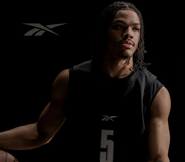 Person wearing a black sleeveless shirt with a Reebok logo, holding a basketball on a dark background.