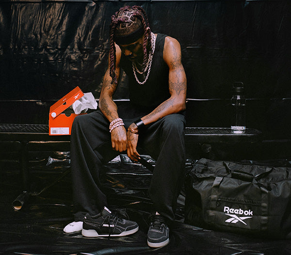 A man sits on a black bench in a dark setting, wearing black athletic pants and black-and-white Reebok sneakers. A black Reebok duffel bag is on the bench beside him, along with an orange shoebox and a water bottle.