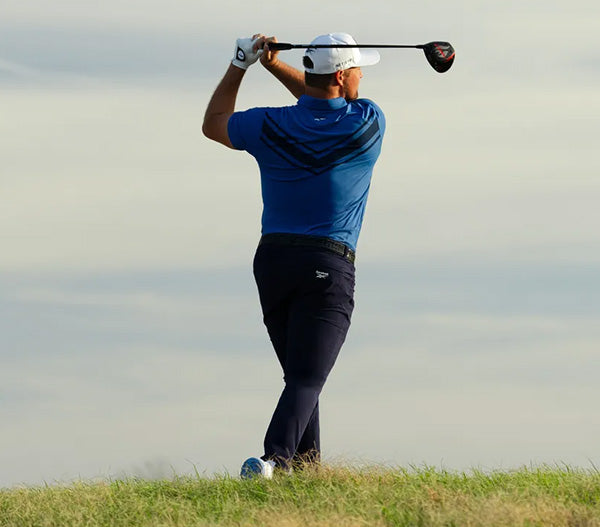 Golfer in blue shirt and black pants swinging a golf club on a grassy field with a clear sky.
