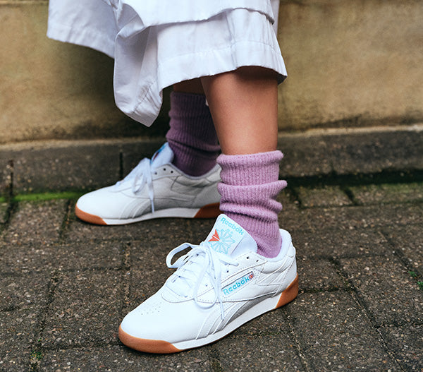 White sneakers with brown soles worn on a person's feet, standing on a pavement.