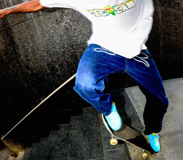 A person skateboarding down dark stone stairs, wearing a white graphic t-shirt, loose blue jeans, and bright turquoise sneakers.