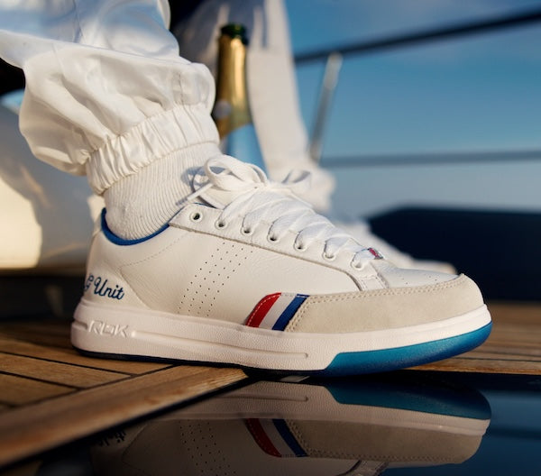A close-up of a white low-top sneaker with blue and red accents, worn with white socks on a wooden deck with a reflection below.